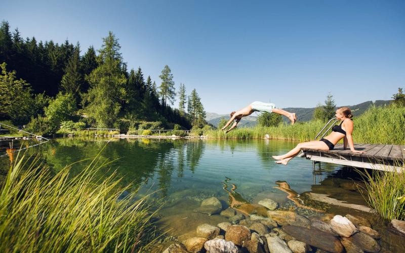 Blick auf den Badeteich mit einer Dame und einem Jungen, der gerade ins Wasser springt, im Hotel Übergossene Alm