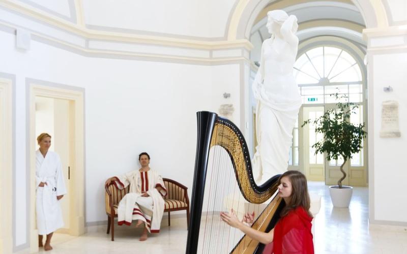 Eine Harfespielerin und zwei Gäste im Bademantel im historischen Teil des Wellnessbereiches im Rimske Terme Hotel Rimski dvor