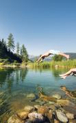 Blick auf den Badeteich mit einer Dame und einem Jungen, der gerade ins Wasser springt, im Hotel Übergossene Alm